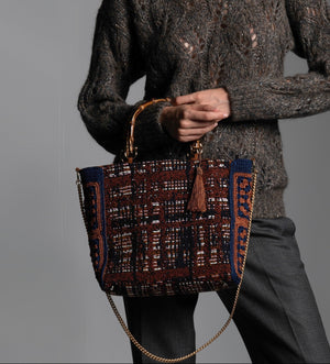 Person wearing a textured knitted sweater holding a patterned handbag featuring bamboo handles, light brown silk thread tassel, and crochet flower side panels.