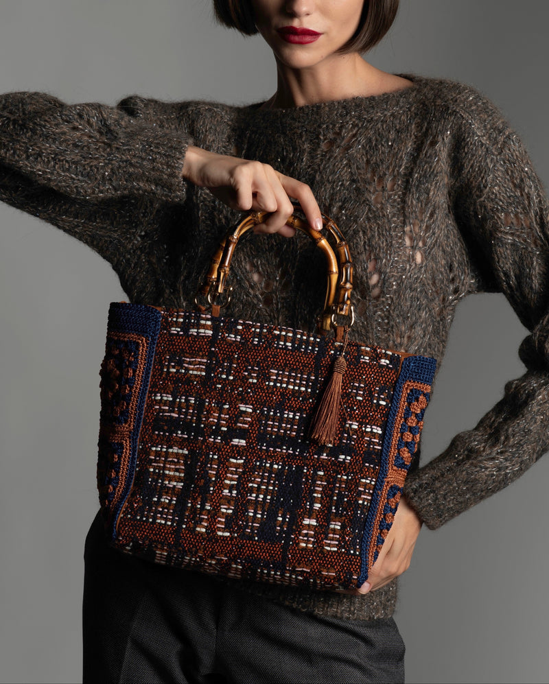 Woman holding a patterned handbag featuring bamboo handles, light brown silk thread tassel, and crochet side panels.  