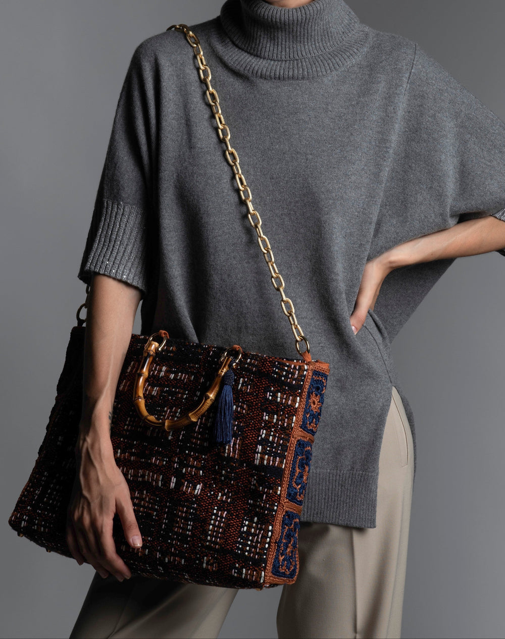 Person wearing a grey sweater holding a patterned handbag featuring bamboo handles, blue silk thread tassel, and diamond cut metal shoulder strap.