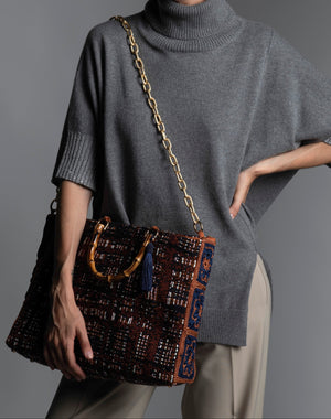 Person wearing a grey sweater holding a patterned handbag featuring bamboo handles, blue silk thread tassel, and diamond cut metal shoulder strap.