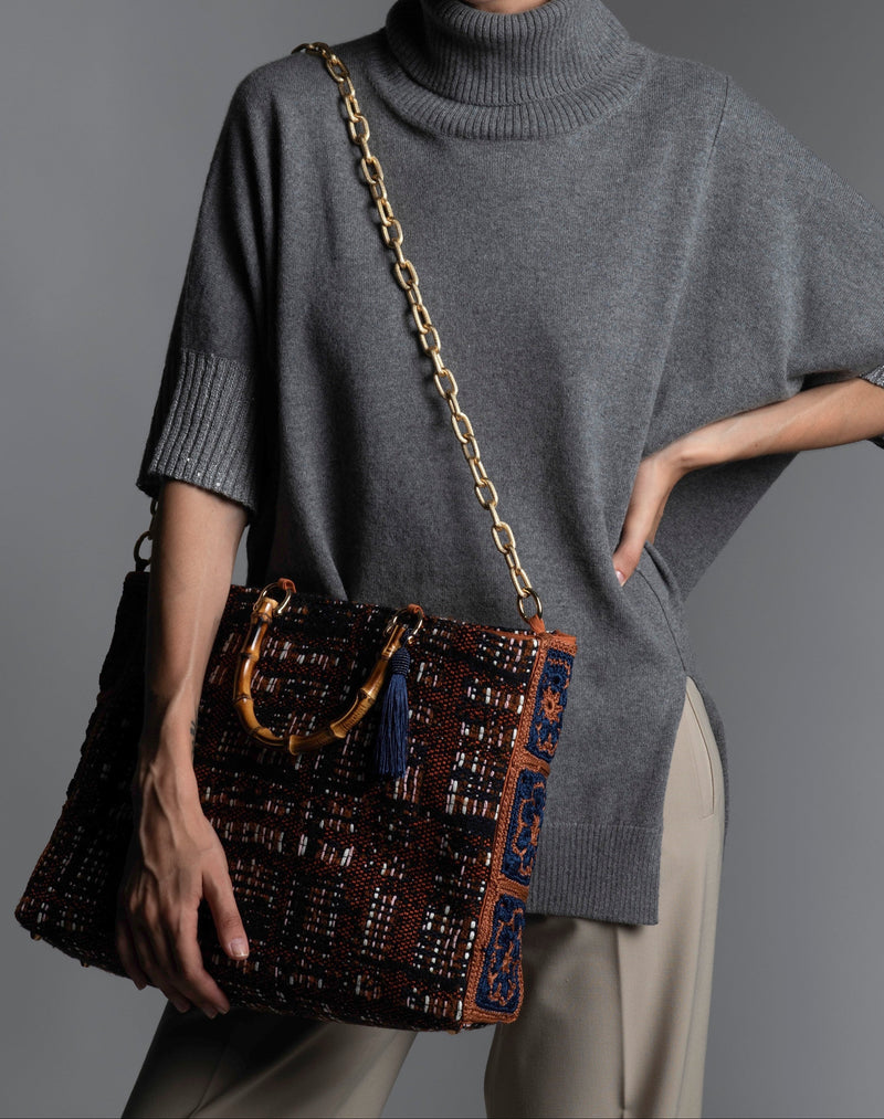 Person wearing a grey sweater holding a patterned handbag featuring bamboo handles, blue silk thread tassel, and diamond cut metal shoulder strap.