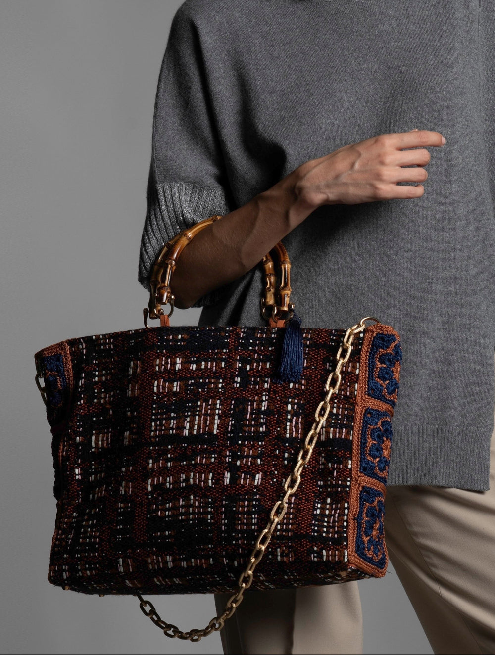Person wearing a grey sweater holding a cotton wool blend handbag featuring bamboo handles, blue silk thread tassel and a diamond cut metal chain. 