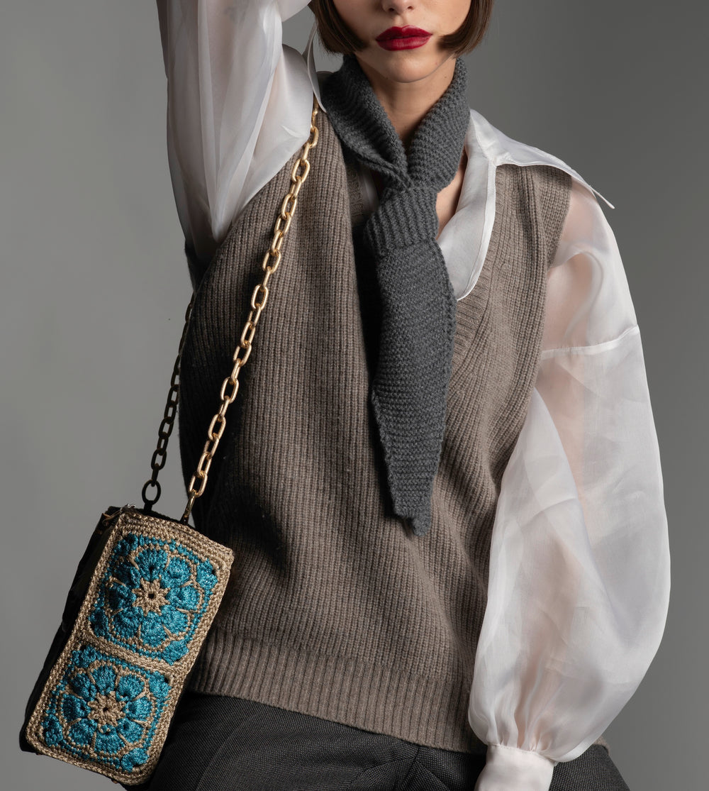 Person wearing a taupe  knitted vest, white organza blouse, and knitted grey scarf, holding a handbag decorated with turquoise crochet flowers.  
