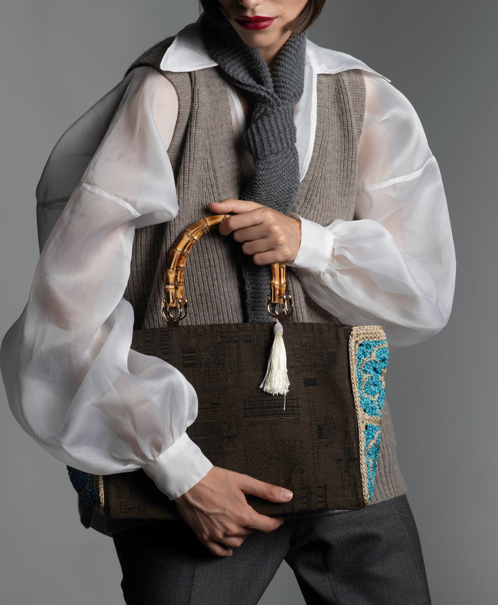 Person wearing a white organza blouse, taupe knitted vest holding a cotton monument-motif handbag featuring bamboo handles and white silk thread tassel.