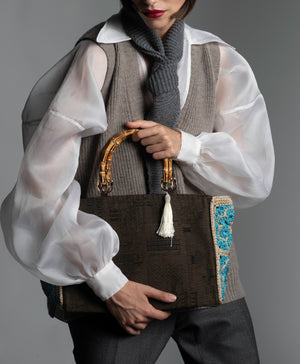 Person wearing a white organza blouse, taupe knitted vest holding a cotton monument-motif handbag featuring bamboo handles and white silk thread tassel.